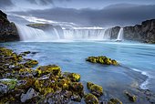 Long time exposure of a turquoise waterfall in volcanic landscape with dramatic clouds and green moss on rocks, Godafoss, Þingeyjarsveit, Norðurland eystra, Iceland