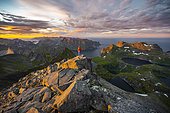 Hiker stands at the summit, sunset with dramatic clouds, view from the top of Hermannsdalstinden, fjords, lakes and mountains, Moskenesøya, Lofoten, Nordland, Norway, Europe