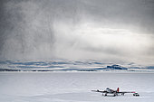 The Basler, stationed on the Terra Nova Bay ice floe in front of the Mario Zucchelli base. The mechanics take advantage of a rare lull to carry out routine maintenance. The aircraft is strapped to the ground. A squall arrives from the left. Victoria Land, Antarctica