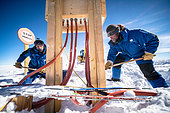 Two technicians finish digging a trench in the snow with shovels, in front of an outdoor electrical box. In summer, a team of around 25 technicians from various trades carry out various maintenance tasks at the resort. This includes the electrical distribution network, which is put to the test by the cold and temperature variations, and whose needs, particularly for scientific installations, are constantly increasing. Manual labor on Concordia is particularly demanding, obviously because of the cold, but also because of the lack of oxygen. Concordia Antarctic Research Station, Dome C plateau, East Antarctica.