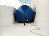 Two technicians install and inflate a cylindrical balloon 5 meters in diameter and 35 meters long. The balloon serves as a mold for the snow to be deposited on top. Once deflated, the result is a cellar spacious enough to store vehicles and other equipment. The beige and orange structure at the far end is the fire escape. Concordia Antarctic Research Station, Dome C plateau, East Antarctica.