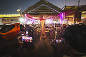 Kumbh Mela 2025: The Sacred Goes Viral. Pilgrims Taking Photos During the Rudrabhishek Ritual. Swamy Gnana Prasanna Giri and Pilgrims Capturing Moments During the Rudrabhishek Ritual. During the sacred Rudrabhishek ritual, which involves the worship of Lord Shiva through chanting and offerings, pilgrims participate not only in the spiritual experience but also in documenting it. The image captures pilgrims holding their smartphones to take selfies and photos, blending the ancient tradition with modern technology. Swamy Gnana Prasanna Giri, affectionately known as 'Guruvu garu' by his followers, is also present, leading the ceremony. The colorful lighting and the energy of the devotees contrast with the deep reverence of the ritual, highlighting how technology has become an integral part of documenting spiritual experiences.