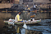 A man selling flower gifts to the Ganges river on his little boat, entirely made by plastic bottleskept together by a fishing net. In the background one of Varanasi's burning ghats.