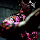 A man wearing a colourful shirt and pushing the camera away
