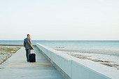 Businessman walking on sidewalk at the beach, pulling suitcase behind him, looking at view